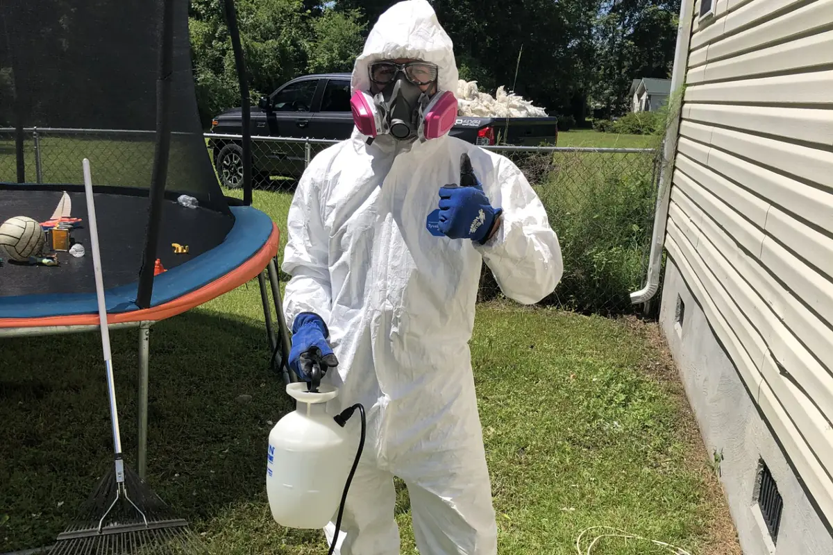 A Bactronix technician in a full protective suit, respirator and gloves, holding a pump sprayer beside a house with foundation vents in the wall behind